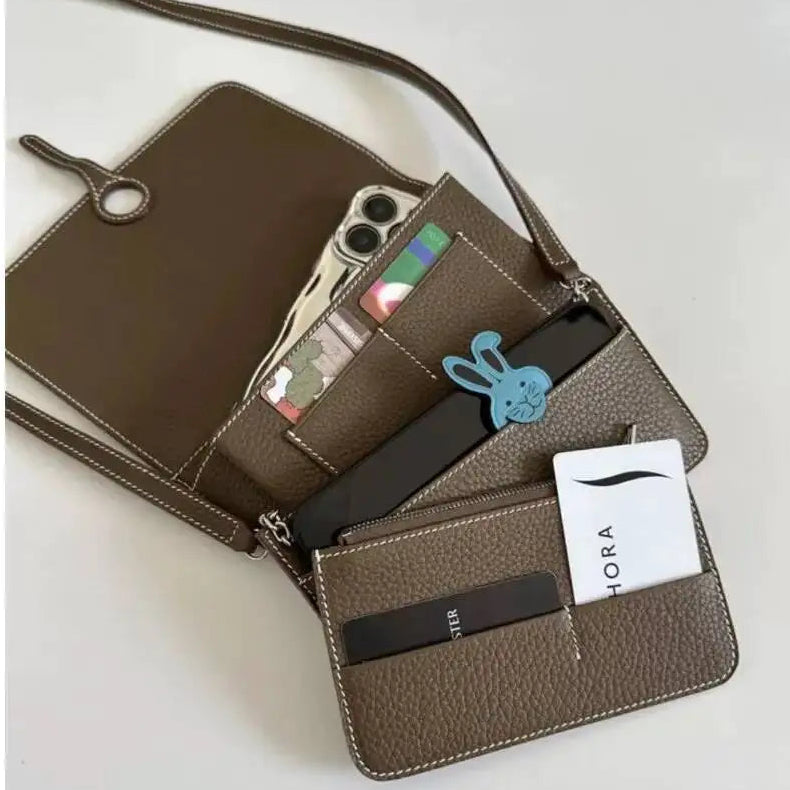 A brown pebbled leather crossbody wallet shown open, displaying its interior card slots, phone pocket, and card holder. The bag is styled with various accessories, including a blue bunny keychain, a black card, and a white HORA card, on a light gray background.