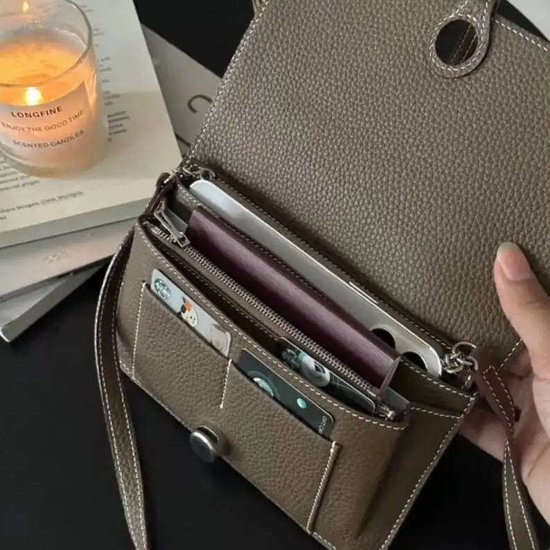 A taupe brown pebbled leather crossbody bag shown open, held by a hand, with its interior compartments, card slots, and zipper visible. The bag is styled on a dark surface next to a lit candle and books.