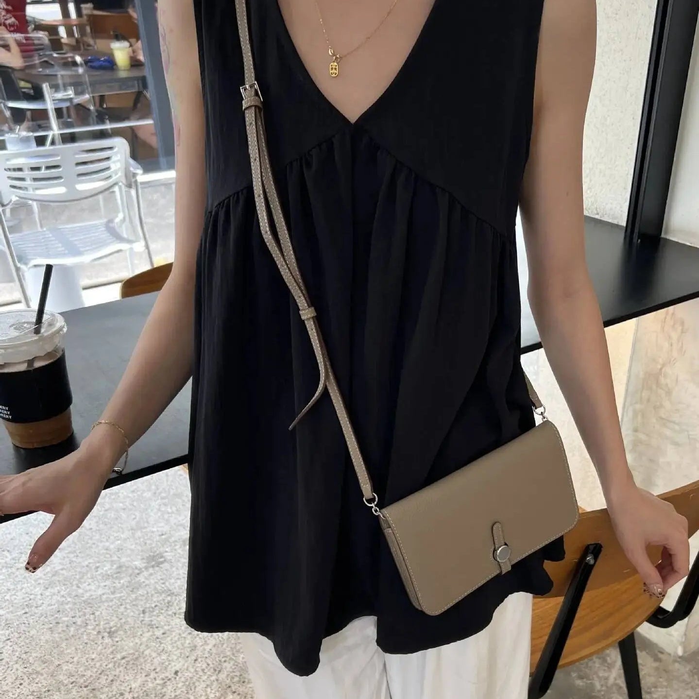 A woman wearing a black sleeveless V-neck top and white pants, carrying a beige crossbody bag with a silver clasp in a cafe setting.
