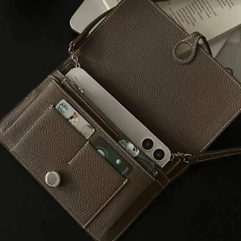A brown pebbled leather crossbody wallet shown open on a dark surface, with card slots holding cards, a phone partially inserted, and a magazine in the background.